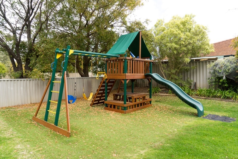 Exterior Playhouse Painting & Staining with Low-VOC Coatings Green and brown wood backyard playhouse with a slide, swings, and climbing ladder on a grassy lawn — exterior playhouse staining and painting Middlesex County MA