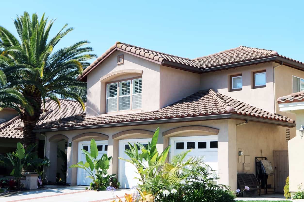 Expert Exterior House Painters Beige stucco home with terracotta tile roof, three white garage doors, and lush tropical landscaping under clear blue sky