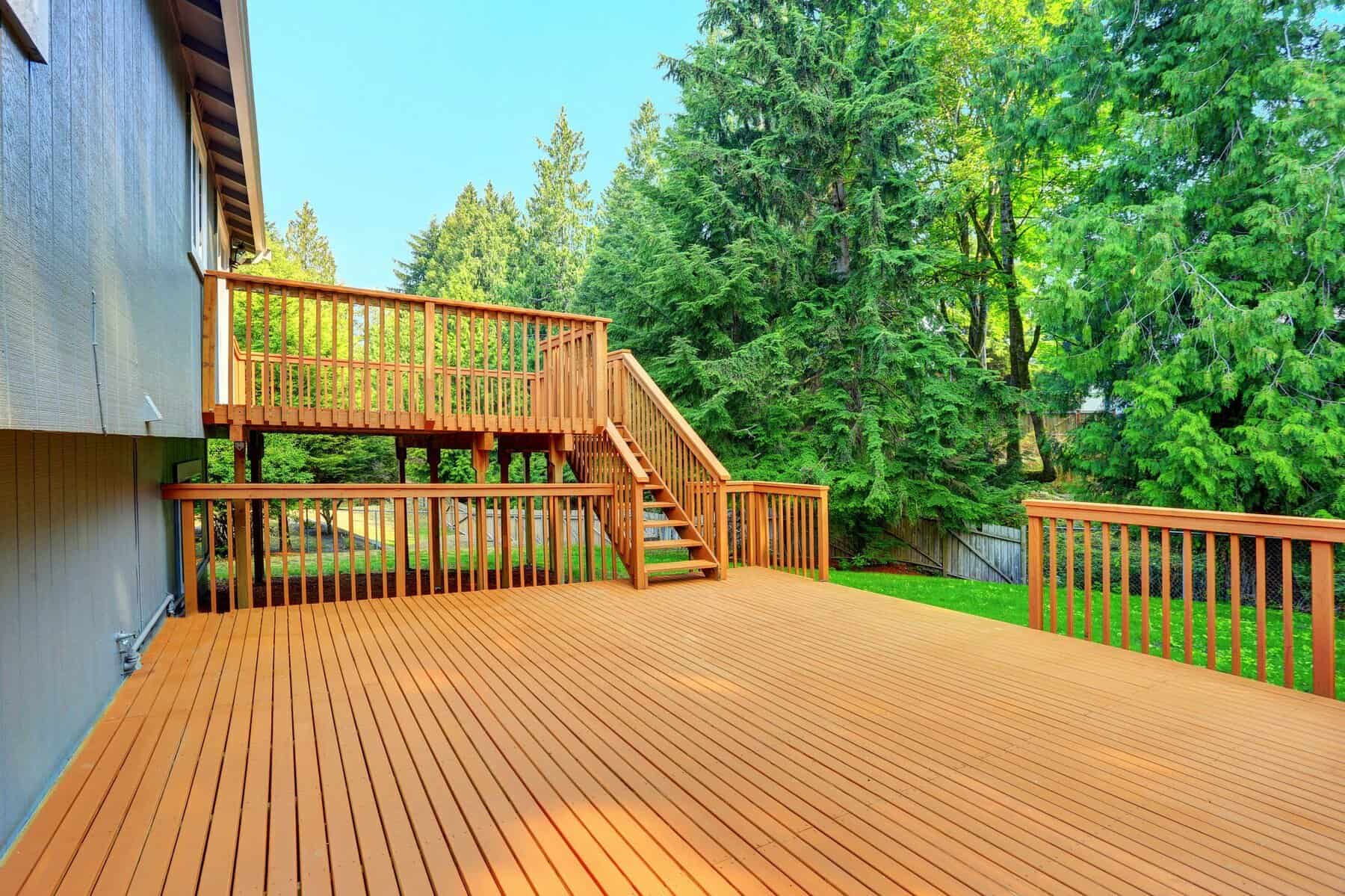 Deck Painting & Staining – Two-Level Wood Deck with Railings Two-level honey-stained cedar deck with railings and stairs beside a blue-gray home — deck painting Massachusetts