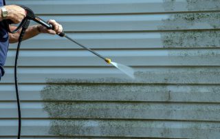Technician pressure washing green mold off blue-gray vinyl siding, revealing a clean streak on a residential home