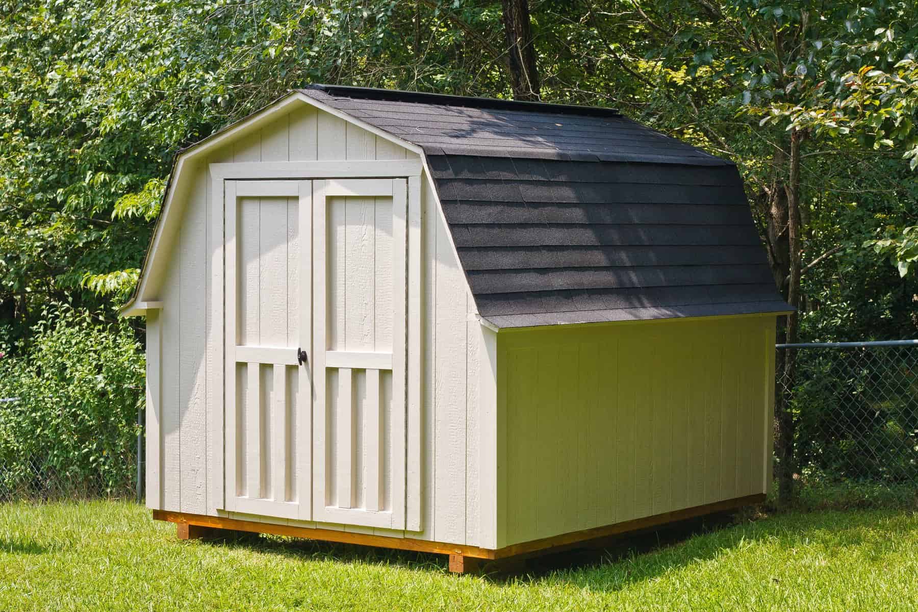 Barn-Style Shed Painting & Staining in Middlesex County MA Freshly painted off-white barn-style shed with double front doors and a dark charcoal gambrel roof, set on green grass near a wooded fence line — shed painting Middlesex County