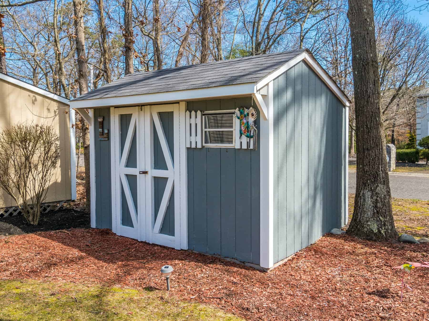 Professional Shed Painting & Staining Services in Middlesex County MA Freshly painted blue-gray wood shed with white trim and double barn doors, nestled among trees on a mulch bed — shed painting Middlesex County MA