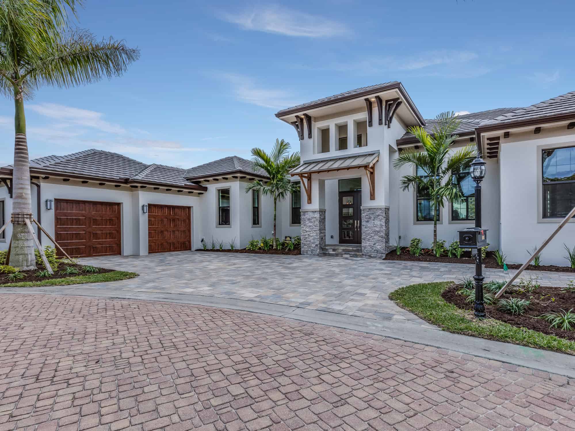 Luxury Home Exterior with Professional Exterior Painting, Stone Accents & Paver Driveway Freshly painted white stucco luxury home with wood garage doors, stone accents, palm trees, and brick paver driveway under blue sky