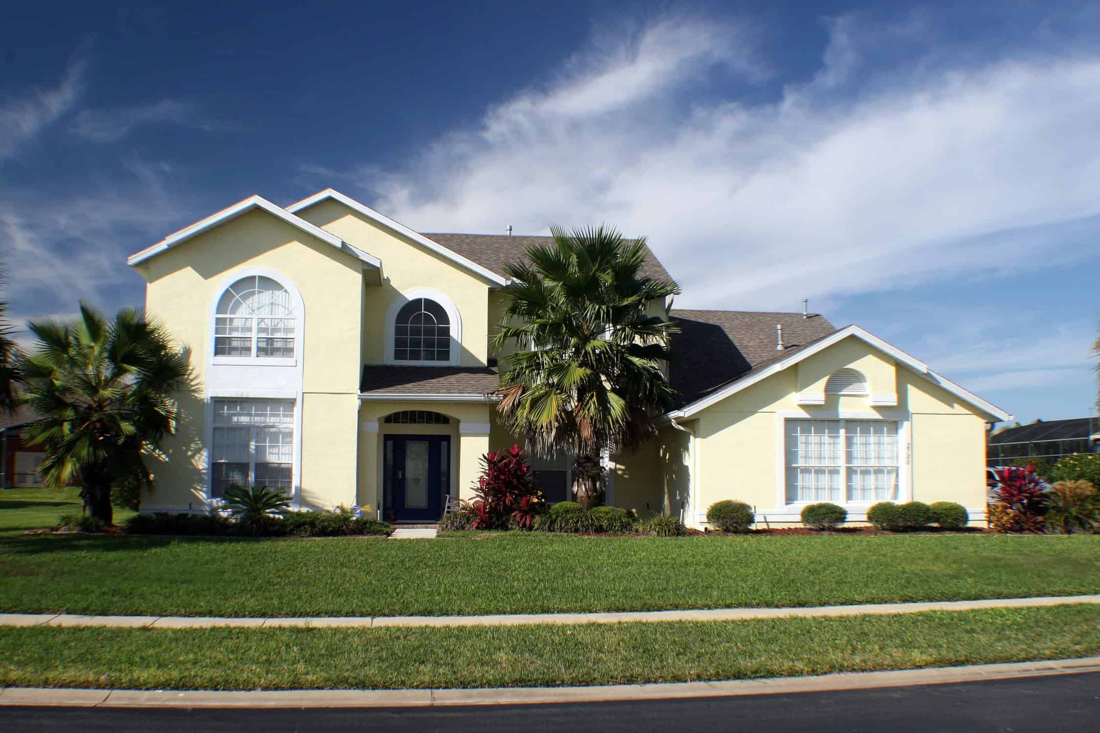 Residential Exterior Painting – Two-Story Stucco Home Light yellow stucco two-story home with navy door, palm trees, and green lawn — exterior painting by licensed house painters.